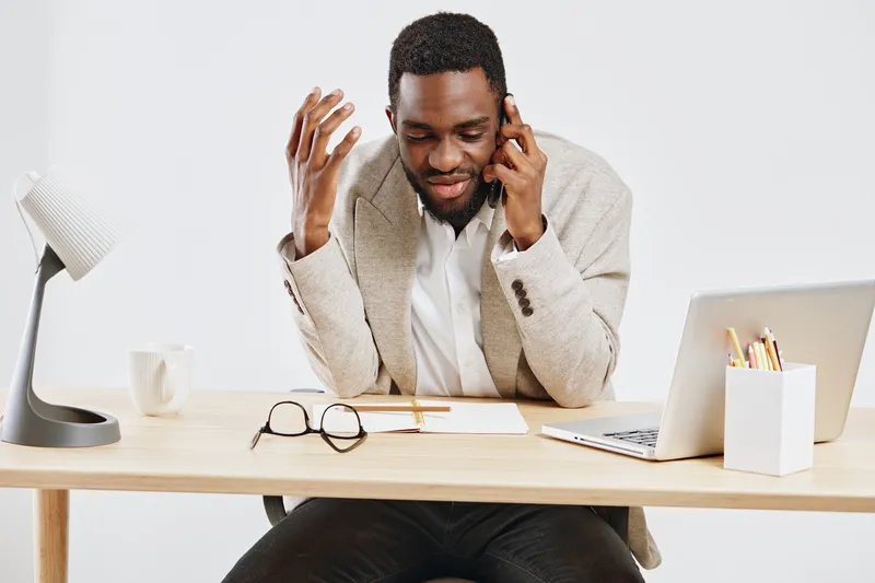 Male African American professional in beige blazer and white shirt sitting at desk using laptop and talking on mobile phone in modern office workspace. Business lifestyle concept