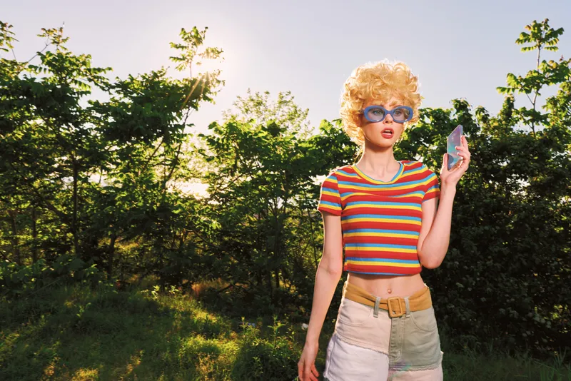 A woman in a striped crop top and light shorts stands outdoors in bright sunlight, wearing retro sunglasses. The scene captures playful summer fashion and carefree lifestyle. Outdoorsy vibe.