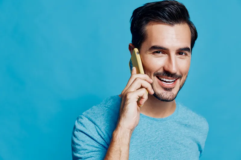 Smiling man talking on mobile phone, wearing light blue shirt, standing against solid blue background, cheerful expression, connecting with loved ones, lifestyle concept.