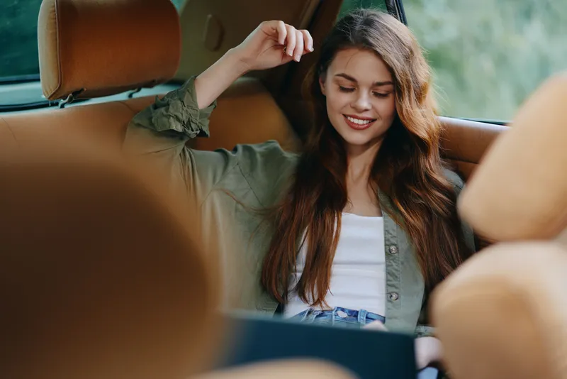 Happy young woman enjoying her time in a car, wearing a casual outfit with long hair, surrounded by a warm, relaxed atmosphere, immersed in her laptop.