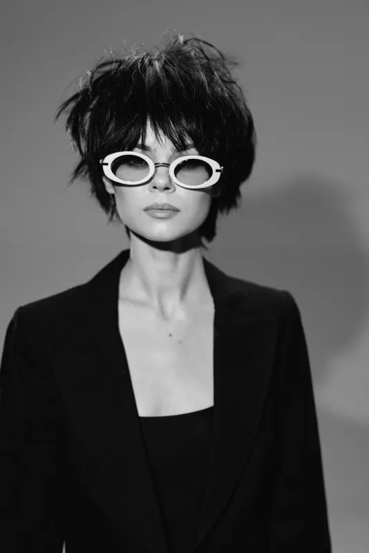 Black and white portrait of a stylish woman wearing white sunglasses and a black blazer with a confident expression.