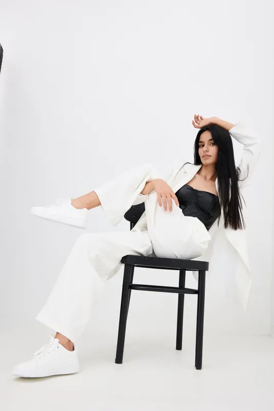 Young woman in white pantsuit and black top posing confidently on black chair against plain white background, wearing white sneakers with relaxed and stylish demeanor.