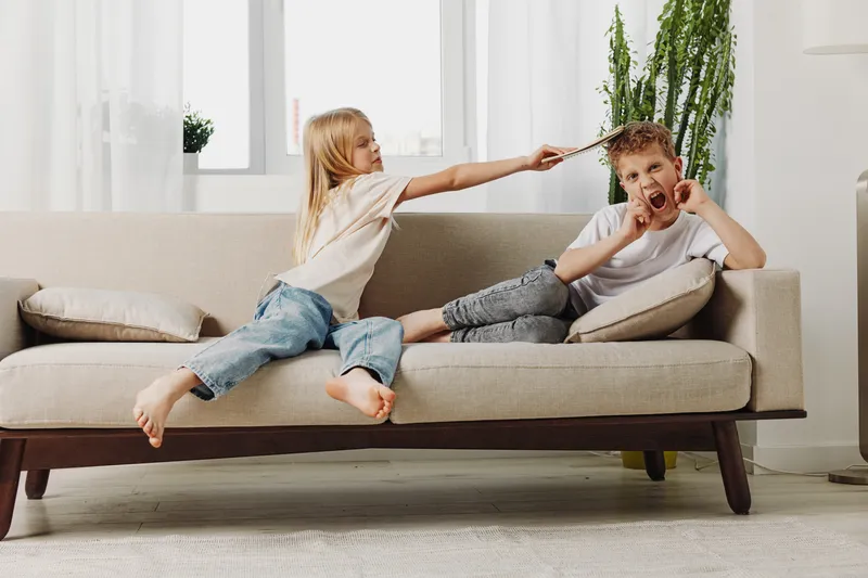 Playful children having fun on beige sofa in bright living room, girl teasing boy with stick, family leisure and sibling interaction concept.
