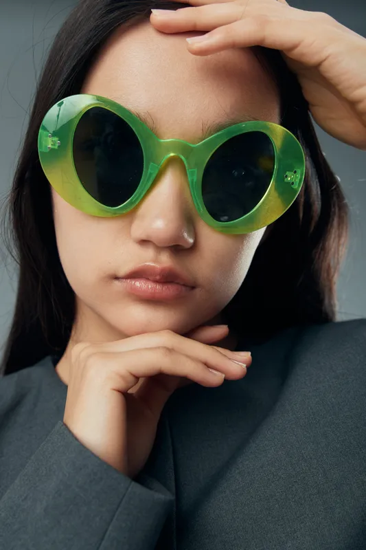 Woman with large green sunglasses and black long hair poses with hands near face in gray outfit on white background. Fashion, style, accessories, beauty, portrait and closeup.