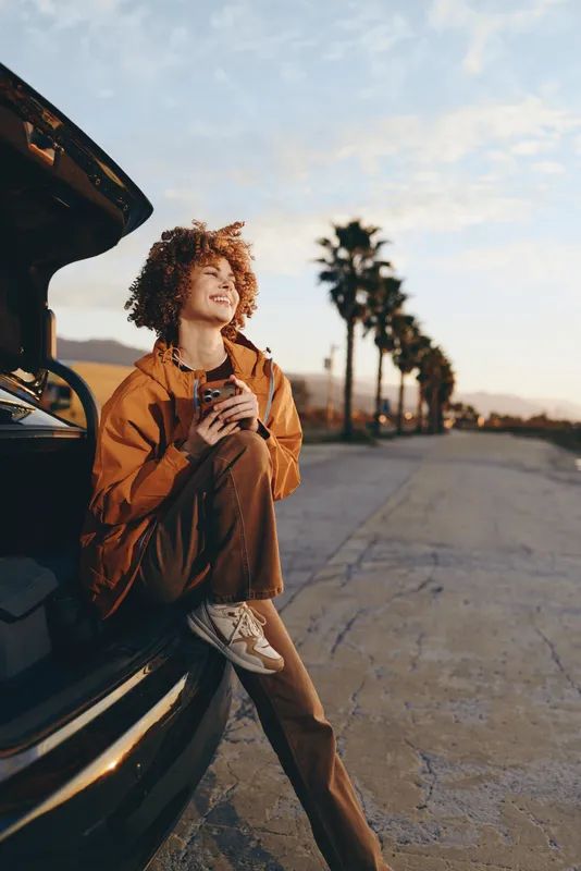 Woman lifestyle outdoors near car at sunset, curly hair smiling and holding coffee, casual jacket, relaxed street mood, soft breeze, warm golden light, palm trees blurred background