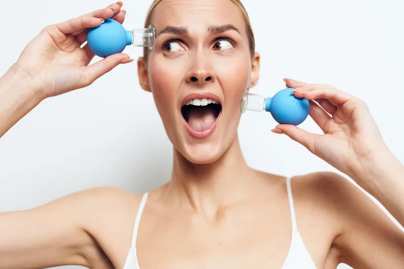 Young woman with surprised expression holding two blue cupping therapy suction cups near her face on white background. Concept of alternative medicine, wellness, and skincare treatment.