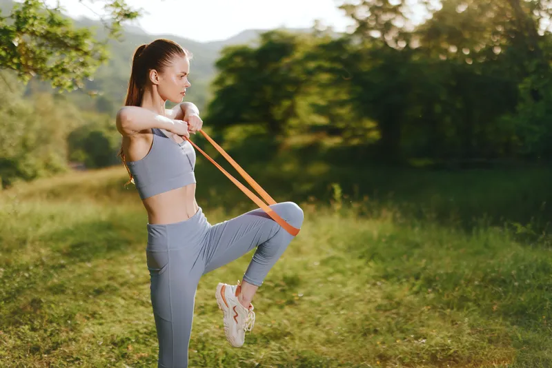 Fit young woman exercising outdoors with resistance bands in sportswear, showcasing strength and determination in a lush green park during summer
