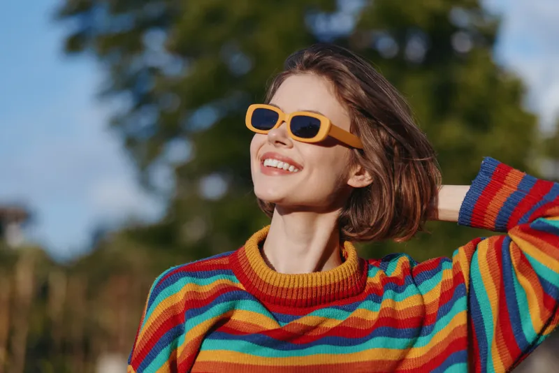 Woman smile sunglasses colorful sweater portrait outdoors, happy fashion young female enjoying sunlight and vibrant style, casual knitted look with positive mood and relaxed confident pose.