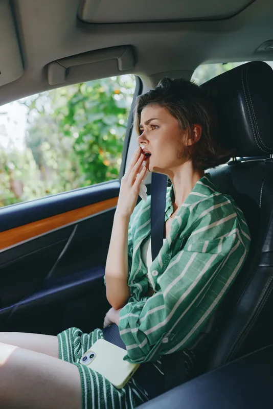 Young woman sits in the front passenger seat wearing a green striped shirt and seatbelt, expressing worry or fatigue while looking toward the window.