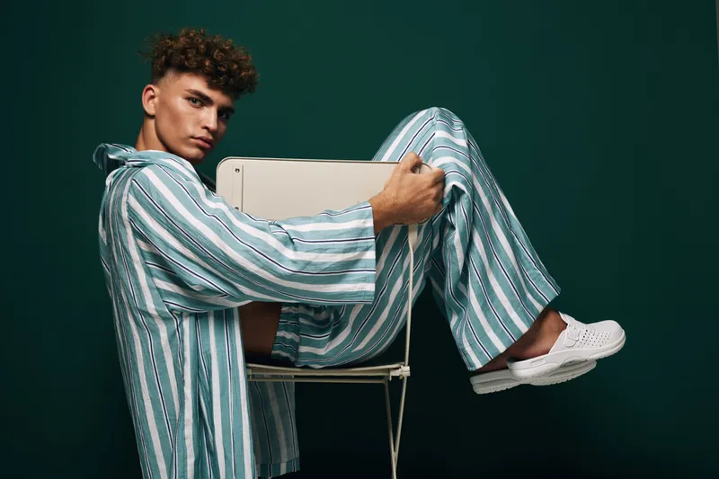 Young man in striped pajamas sitting on chair with laptop on dark background. Casual outfit, technology, and relaxed pose for work or study at home or studio.