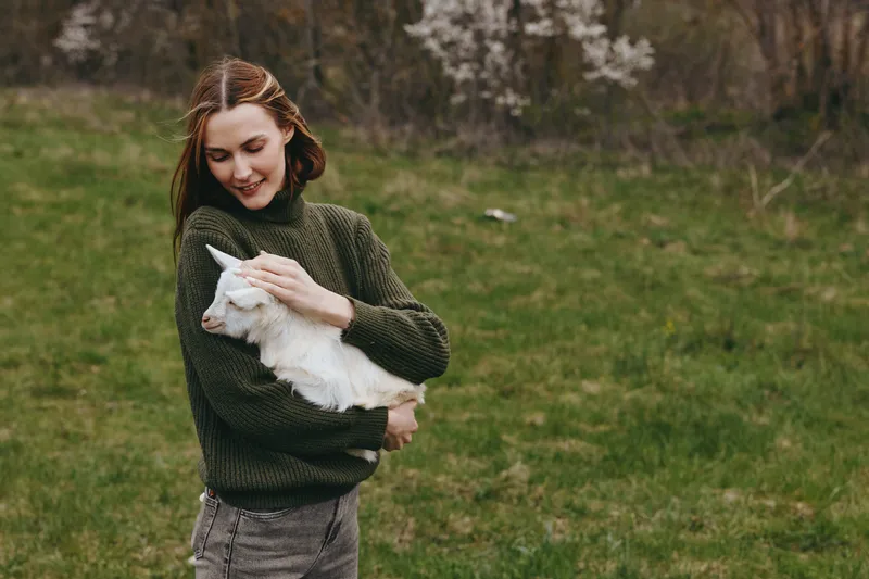Lifestyle outdoor moment with a woman holding a goat kid in a green sweater, smiling softly, sunlit field, calm breeze, natural colors, lush meadow, affectionate interaction, simple countryside charm