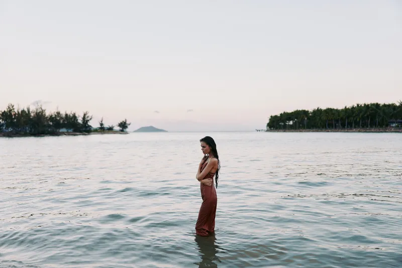 Woman stands waist-deep in calm sea, wearing a red dress, gazing toward the horizon as distant islands and trees frame a serene coastal scene.