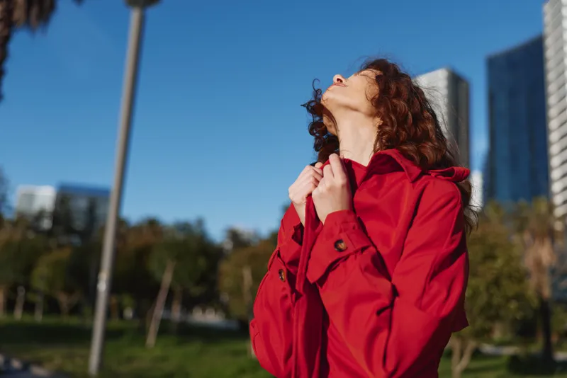 Young woman with curly hair wearing a red coat looks upward while standing in an urban park on a sunny day with modern buildings in the background.