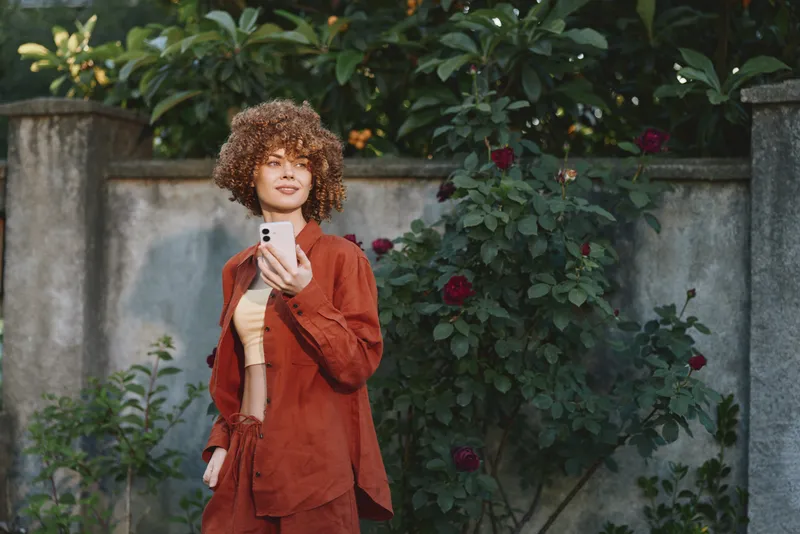 Young woman with curly hair wearing a rust jacket stands outdoors by a flower wall, looking at her phone, a candid lifestyle moment