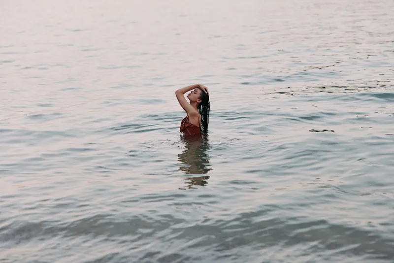 A lone swimmer stands waist deep in calm water, hands through hair, serene pose as soft light glints on gentle waves, capturing a peaceful, intimate bath by the sea.