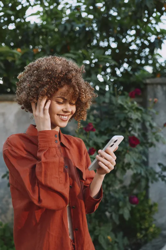 Young woman with curly hair in an orange jacket smiles while checking her phone in a garden setting, feeling relaxed and connected