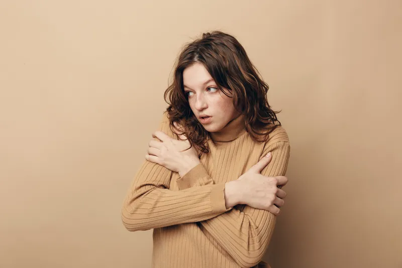 Young woman in beige sweater hugs herself with crossed arms, looking away with worried expression. Neutral background highlights emotion of anxiety and discomfort.