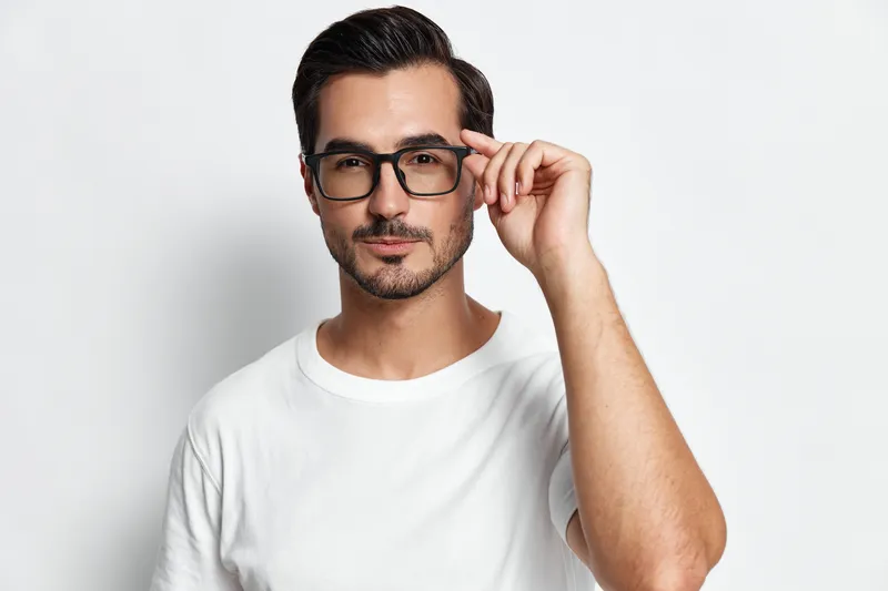 Stylish young man adjusting his glasses while looking confidently at the camera, wearing a casual white t-shirt against a clean grey background. Modern lifestyle theme.
