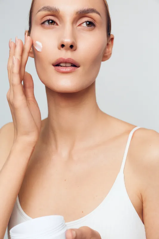 A young woman in a white tank top applies cream to her cheek with her fingers in a bright studio. The image represents skincare, beauty routine, and healthy skin care concepts.