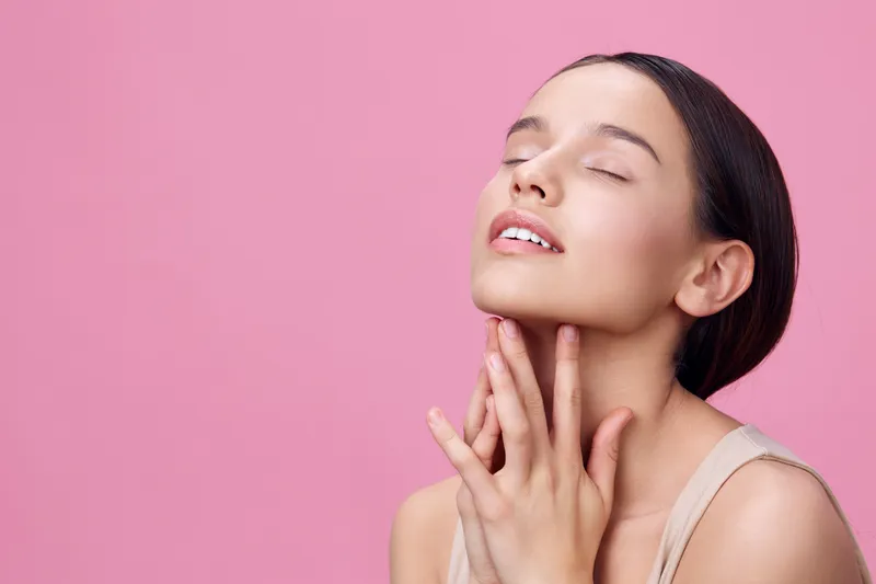 Young woman with closed eyes and serene expression, touching her face, isolated on pink background, conveying beauty and self care concepts in a minimalist style