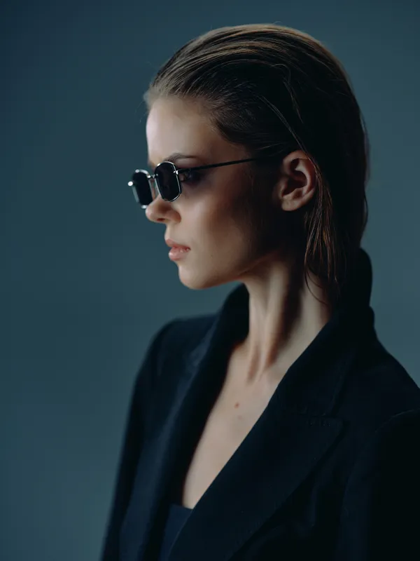 Stylish woman wearing black sunglasses and a dark jacket with a serious expression against a dark blue background.