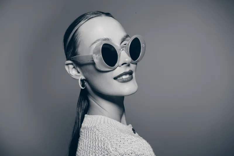 Portrait of a young white woman with light skin wearing large round sunglasses and a knitted sweater. She poses confidently with slicked back hair in monochrome studio lighting.