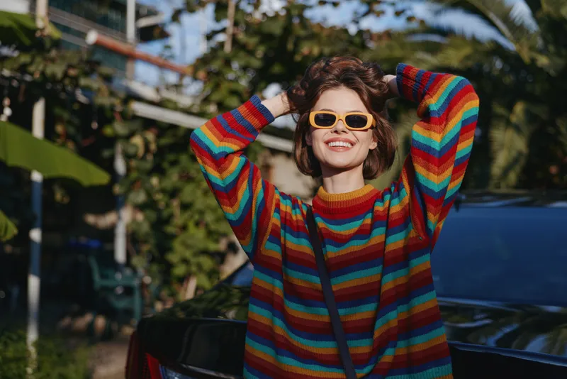 Woman in a colorful striped sweater and yellow sunglasses smiling with hands behind head beside a car in tropical outdoor setting, young adult casual outfit for relaxed travel lifestyle.