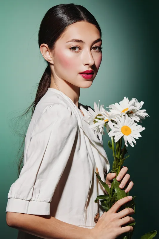 Young woman with elegant makeup holds white daisies in her hand wearing a soft white shirt. Studio portrait with clear skin and delicate beauty fashion concept.