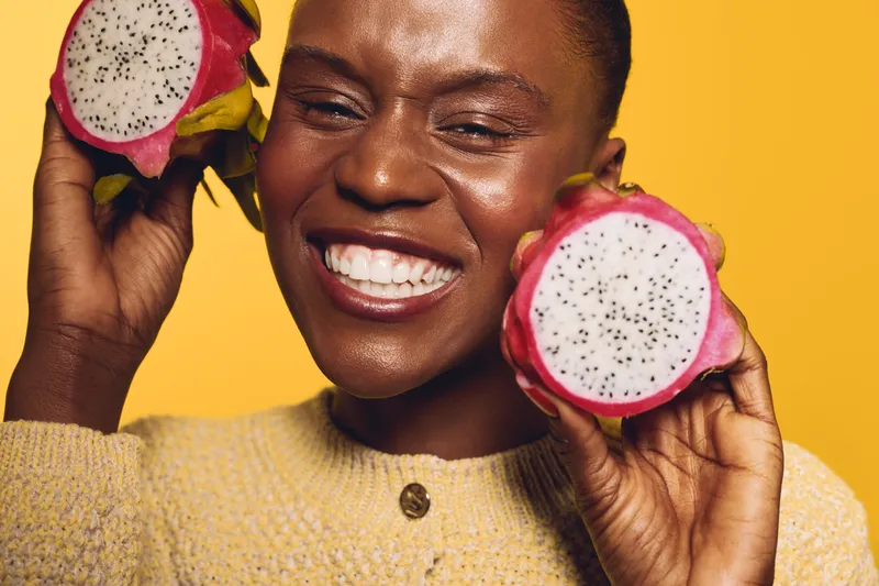 Happy African woman with dark skin holding two halves of dragon fruit near her face on yellow background. Concept of healthy lifestyle, fruit, nutrition, and natural beauty.