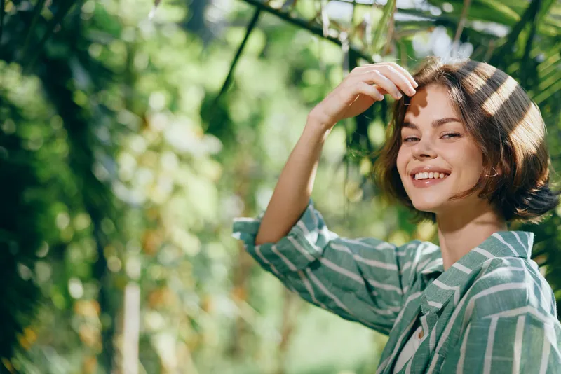 woman smiling in an outdoor portrait, a cheerful gaze framed by green foliage and warm sunlight, conveying natural happiness, relaxed style, friendly energy, and serene outdoor atmosphere for a fresh