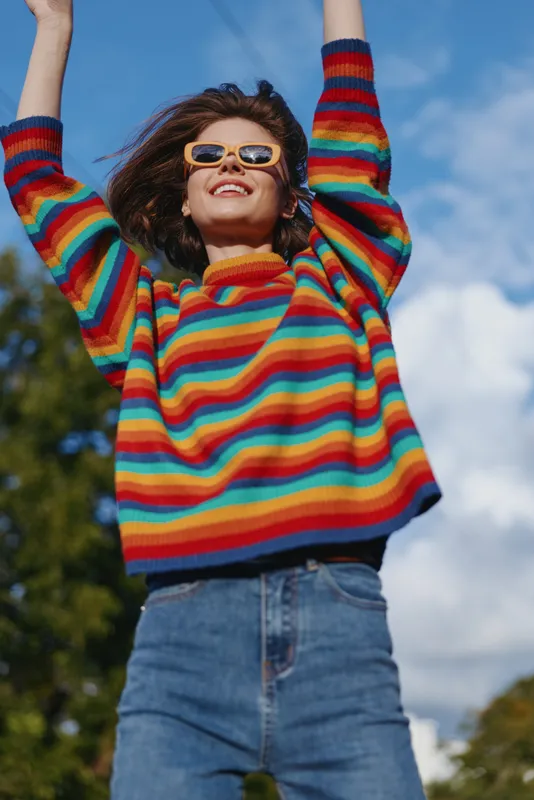 Woman smiling in a rainbow sweater and sunglasses with arms raised outdoor against blue sky; young adult in jeans, casual outfit, joyful expression for lifestyle and fashion travel.