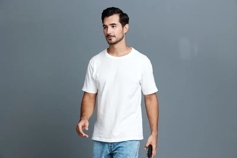 Casual smiling man wearing white t-shirt and jeans holding smartphone while walking confidently towards the camera on solid grey background. Lifestyle concept