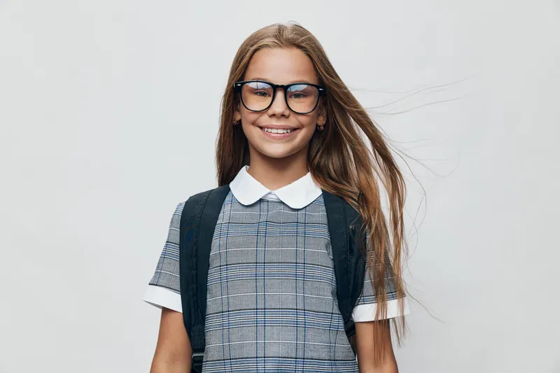 Girl with long hair and glasses smiling wearing school uniform and backpack standing against gray background looking confident and happy.