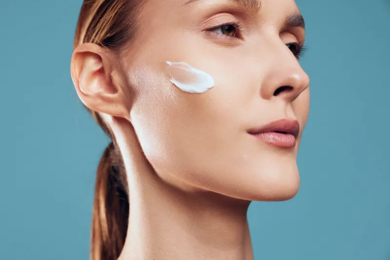 Close up portrait of a young adult woman applying facial cream to her cheek in a clean studio, conveying skincare routine and beauty focus.