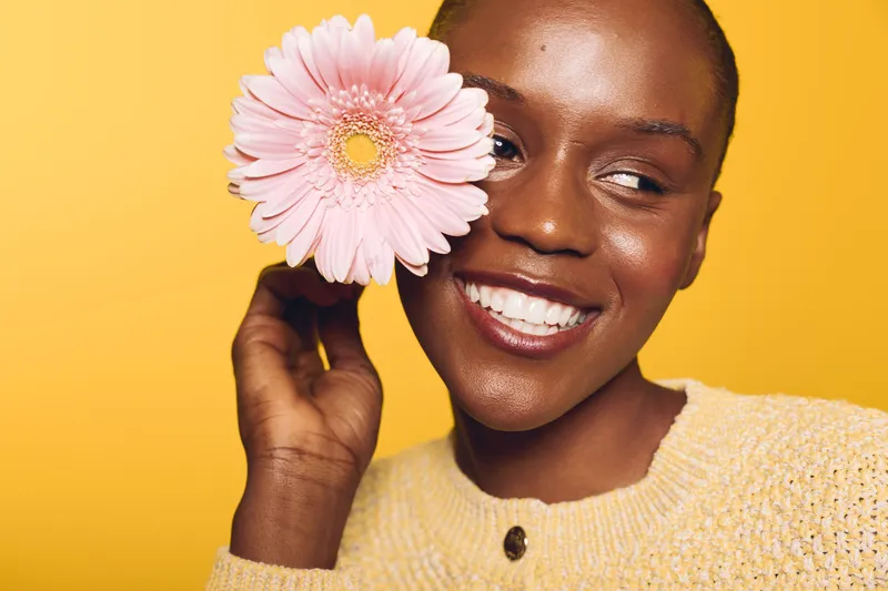 Smiling dark skinned African woman holds pink flower near face on yellow background. Concept of beauty, happiness, natural skin, and floral freshness in studio portrait.