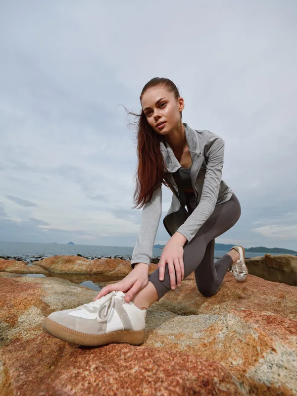 Athletic young model in sport attire stretching on rocky beach during cloudy day, showcasing healthy lifestyle, fitness, and determination