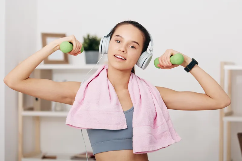 Woman fitness sport enthusiast performing a workout with headphones, dumbbells and a towel draped over shoulders in a bright indoor gym, conveying health, strength and motivation.