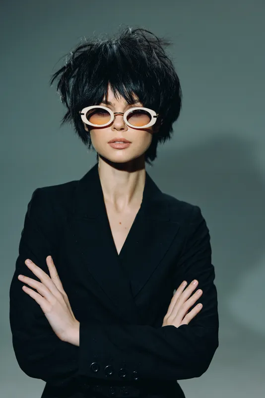 Stylish woman with short black hair and unique sunglasses wearing a black blazer with arms crossed against a plain gray background.
