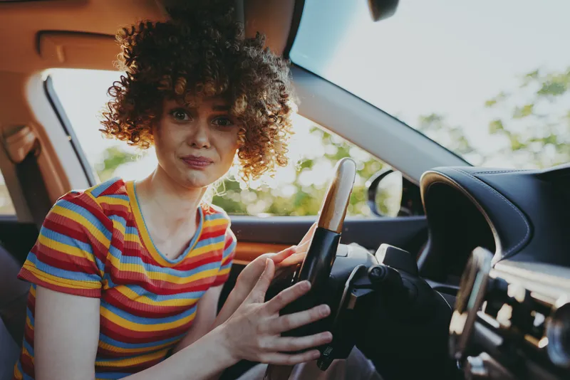 Woman driver with curly hair sits in a car, wearing a colorful striped shirt, capturing a casual lifestyle moment with retro film tones and warm, candid charm.