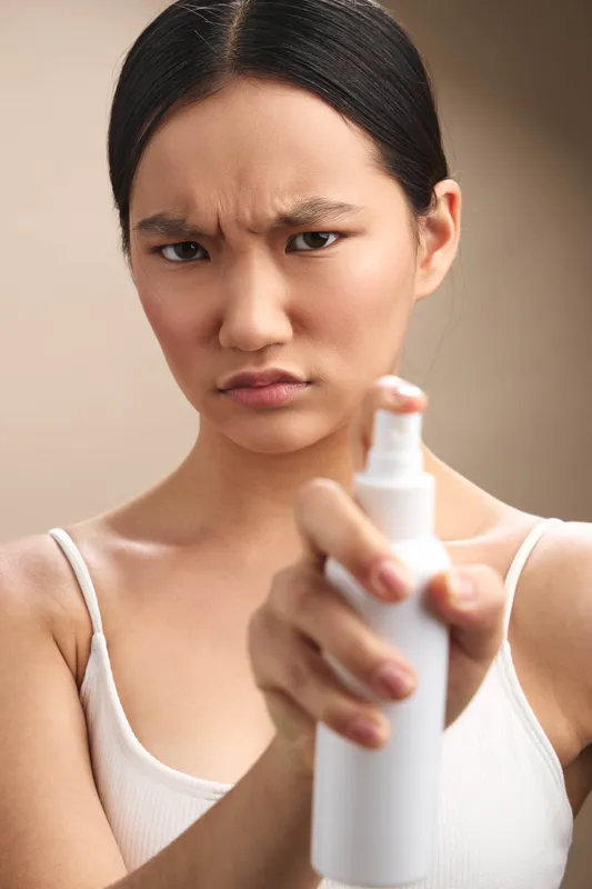 asian woman with a tense expression aims a spray bottle toward the camera, conveying focus and urgency in a neutral setting, suggesting preparation, caution, and personal care.