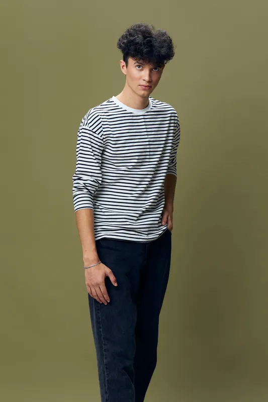 young man wearing striped shirt and dark pants standing against olive background, casual style, natural expression, contemporary fashion portrait