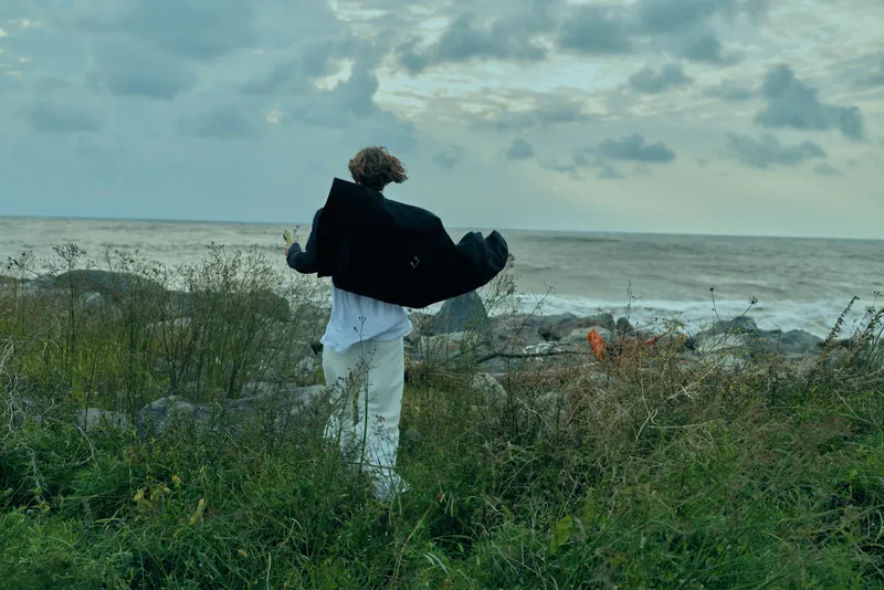 Windy coastal scene featuring a figure in white pants and a flowing dark coat swept by the breeze, standing near the shoreline with sea spray, moody clouds and an open horizon