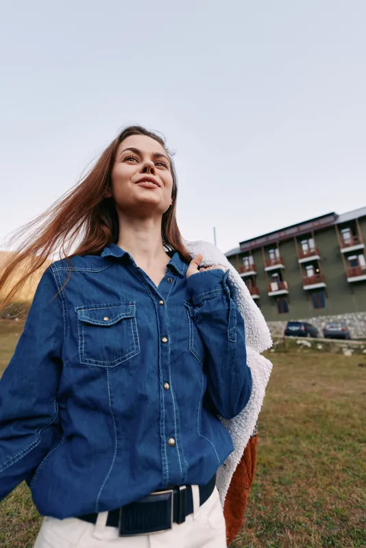 Woman portrait denim outdoors smile, wearing denim shirt and carrying jacket over shoulder near a hotel, casual travel lifestyle with low-angle candid pose and soft natural light