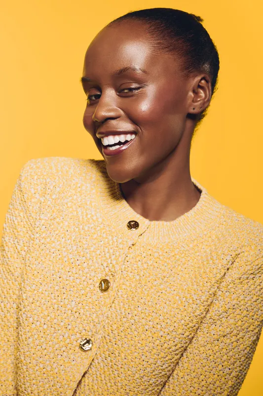 Portrait of a smiling dark skinned African woman with shiny skin wearing a yellow textured jacket on a yellow background. Concept of beauty, happiness, and natural skincare.