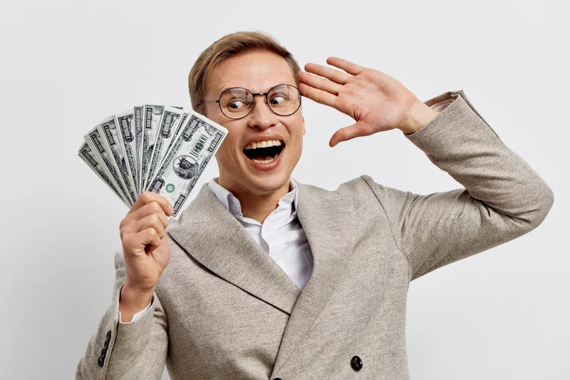 Cheerful middle-aged man wearing glasses and a beige suit celebrating success with a big smile, holding dollar bills, isolated on a plain white background. Business achievement concept.