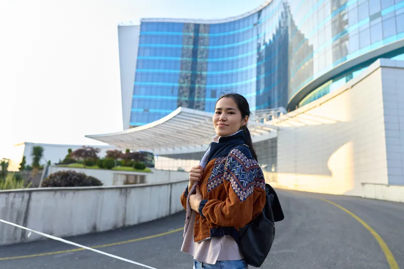 Woman with backpack in a stylish scarf standing beside a modern glass building in an outdoor city scene, casual fashion and confident pose, daylight, architectural backdrop, street photography vibe