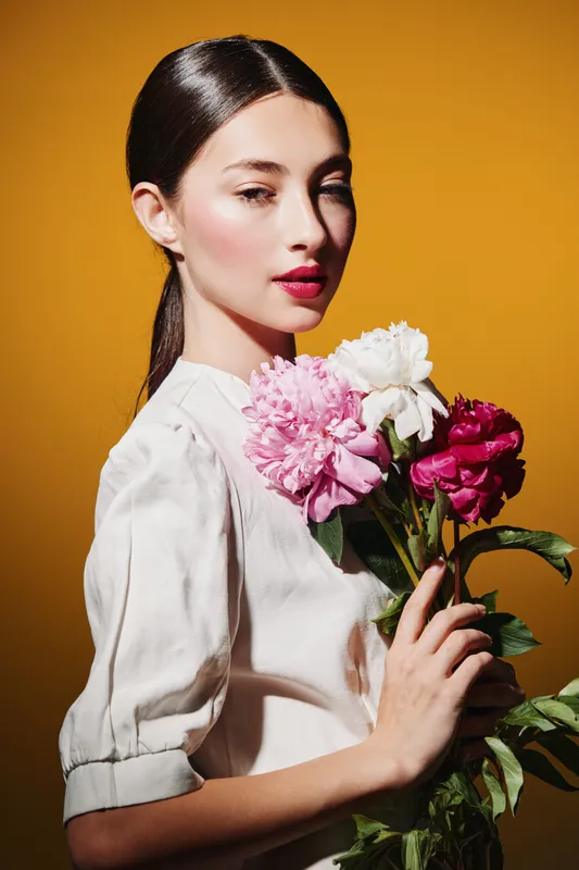 Elegant woman with sleek hair holds vibrant pink and white flowers against orange background. Close-up portrait for beauty fashion and creative floral concept.