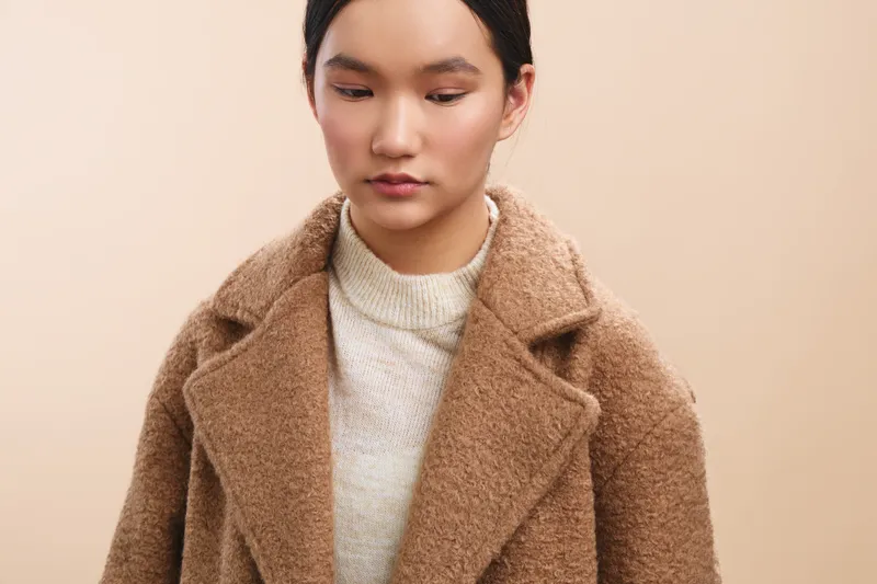 asian woman wearing a tan wool coat stands against a soft beige background, calm and poised, presenting a stylish fashion portrait with warm, understated mood.