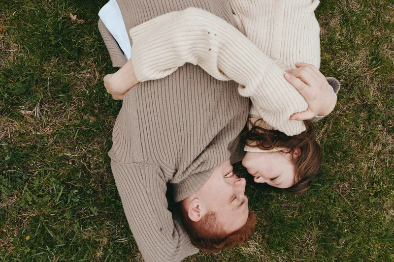 A couple lies on grass in cozy sweaters, facing each other with smiles, bathed in natural light. The scene captures intimate connection, relaxed mood, and a casual outdoor lifestyle between two