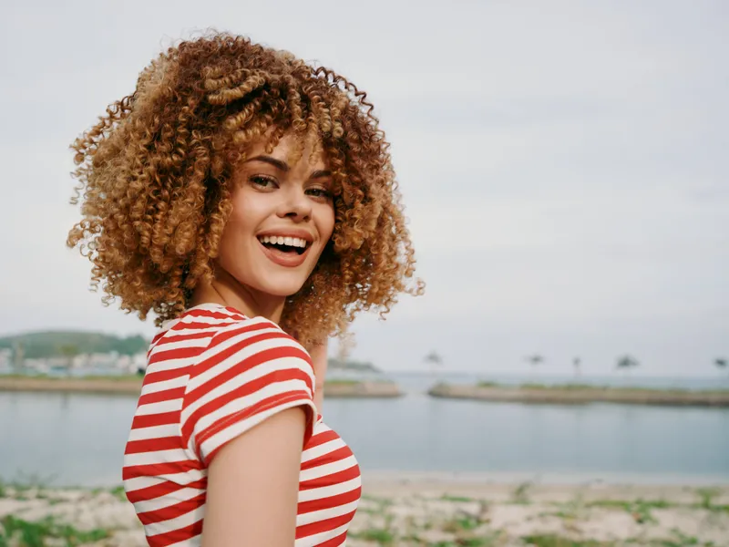 Cheerful young woman with curly hair smiles at the camera by a tranquil waterfront, wearing a red and white striped top, bathed in soft natural light, exuding warm summer happiness.